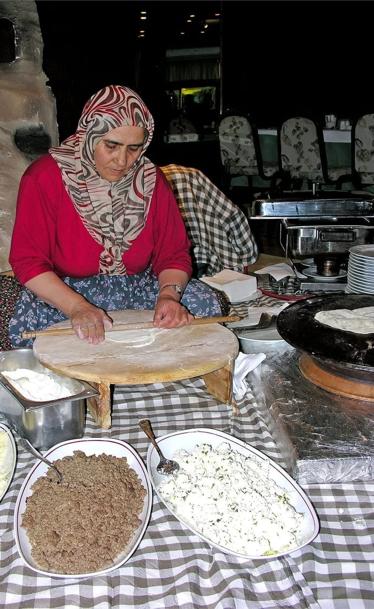 A village cook rolling gözleme dough by hand at a low table beside a hot iron griddle