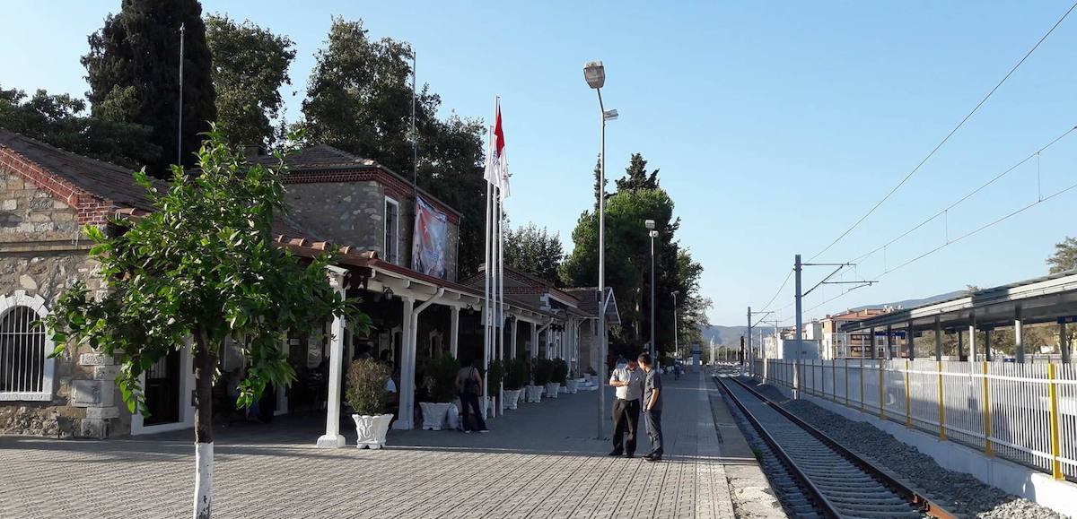 The old stone railway station at Selçuk with tiled roofs, a covered porch and a Turkish flag, beside a paved platform and an electrified track running into the distance