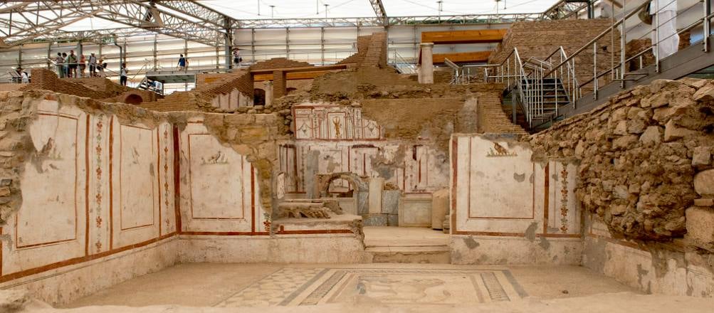 A room in the Terrace Houses of Ephesus with red-paneled wall frescoes and a geometric mosaic floor under the modern shelter