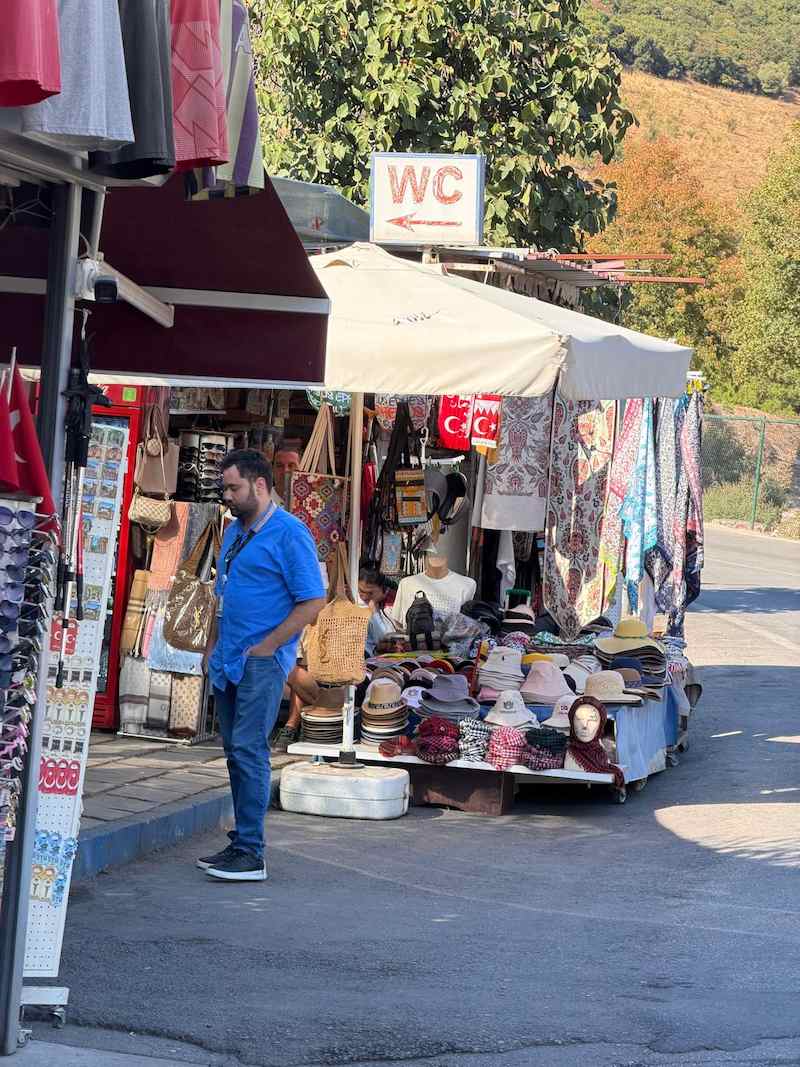 A WC sign with a red arrow above souvenir stalls outside the upper gate of Ephesus, with racks of sun hats, scarves and bags under a white umbrella and a man in a blue shirt standing in front