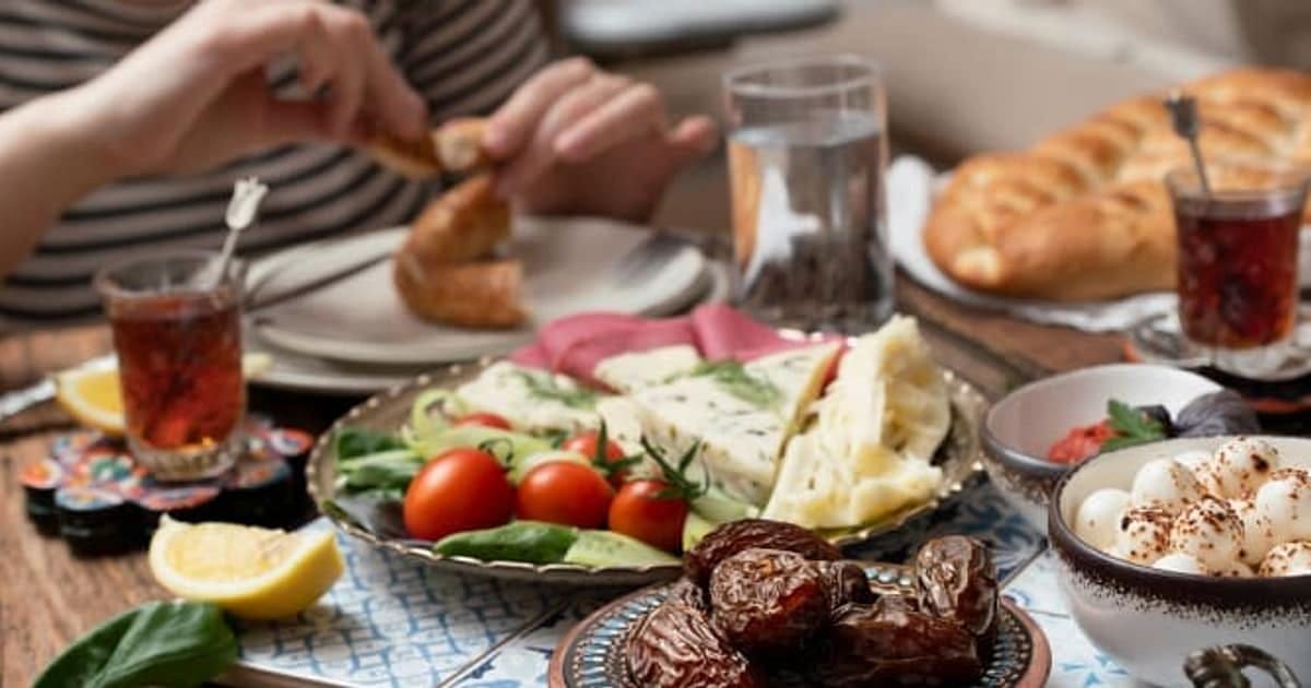 Hands breaking bread over a laid Turkish table of cheese, tomatoes, olives, dates and tea